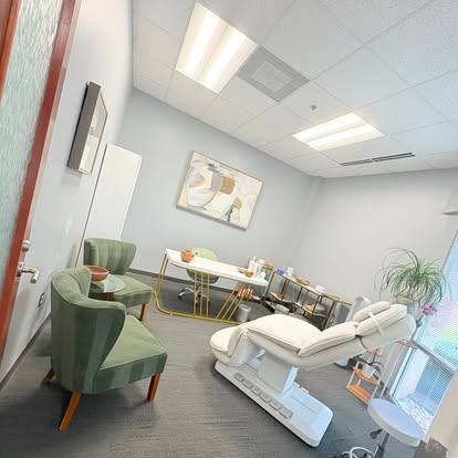 A tidy spa treatment room with a white treatment bed, green armchairs and a gray carpet
