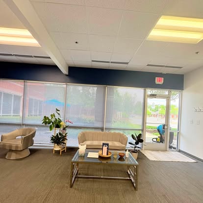 An office waiting area with a beige sofa, an armchair and a glass coffee table in front of tall windows